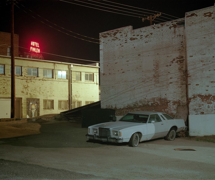 A white car is parked next to a brick building with a red neon sign that reads "Hotel Finlen" at night.