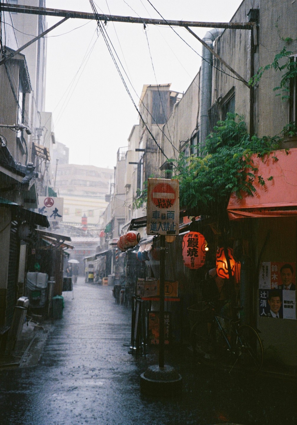 A narrow street in Japan is wet from the rain, with buildings and shops lining both sides and a person with an umbrella in the distance.