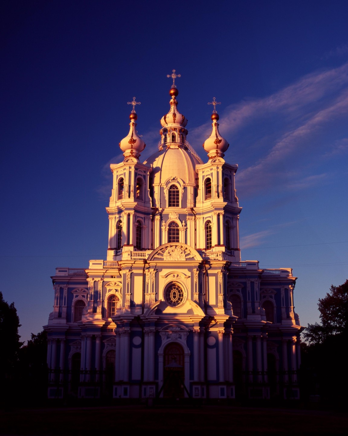 A grand, ornate cathedral with multiple domes and crosses is illuminated by golden light against a deep blue sky.