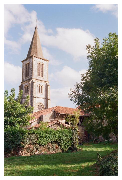 A stone church with a tall steeple stands behind a building with a red tile roof and a green hedge.