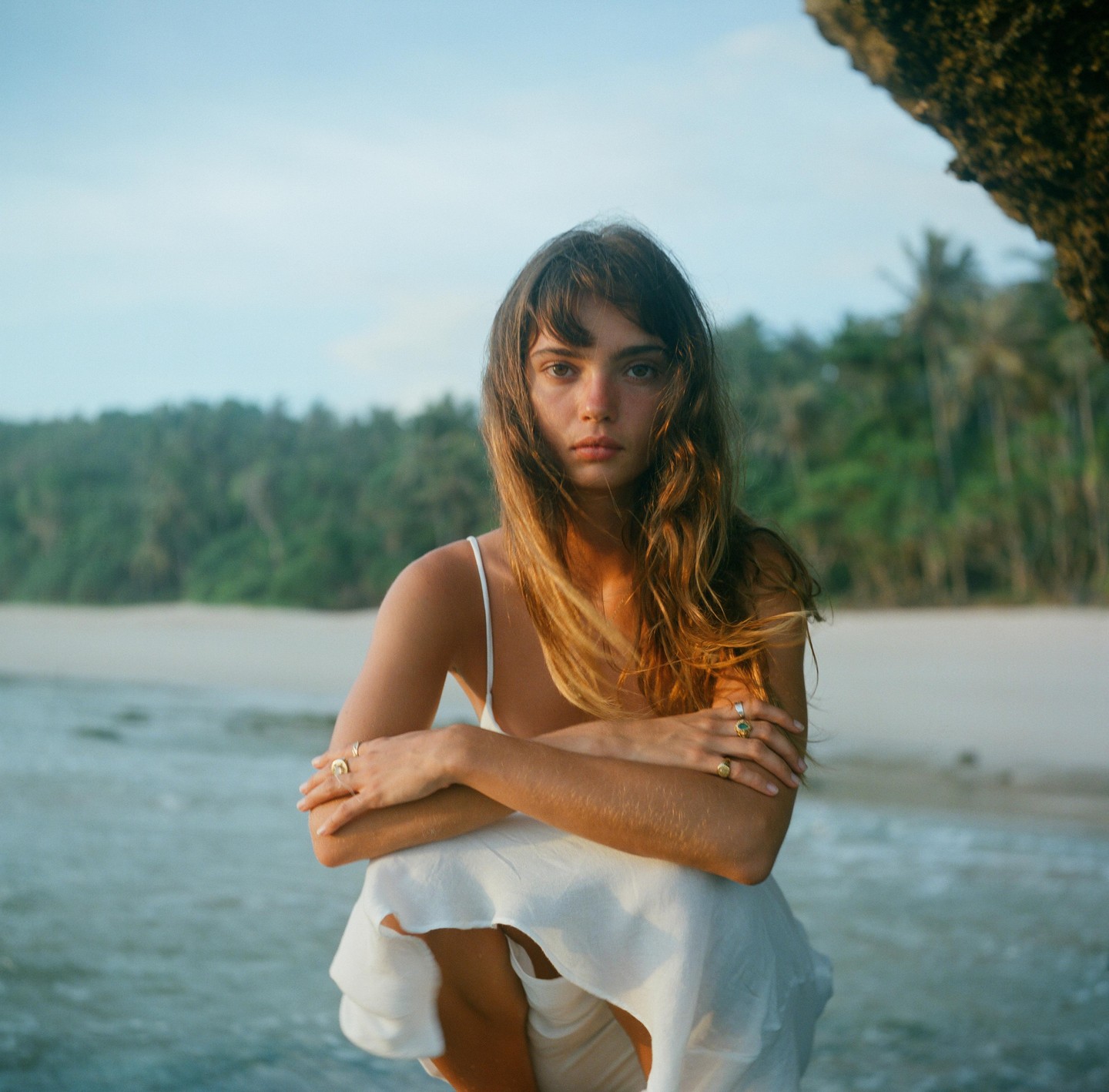 A woman with long hair and a white dress crouches by the water on a beach with a forest in the background.
