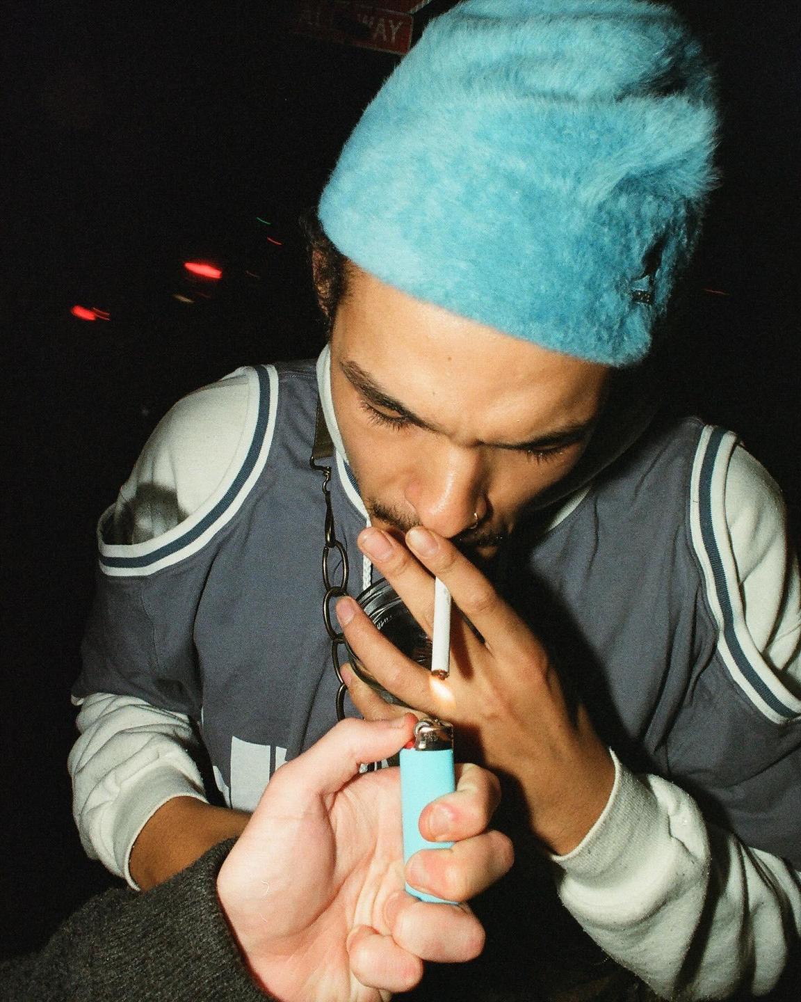 A man in a blue hat lights a cigarette held in his mouth.