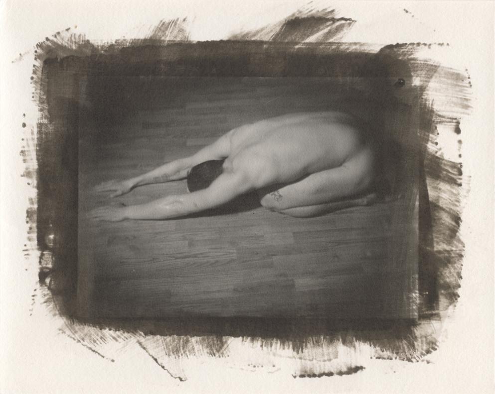 A nude man is kneeling on a wooden floor with his head down and arms stretched forward.