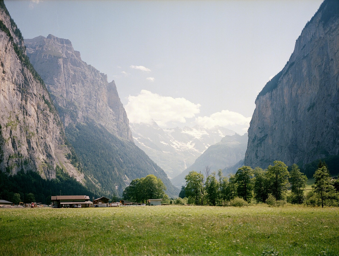 A valley with a green field and buildings is surrounded by tall mountains under a bright sky.