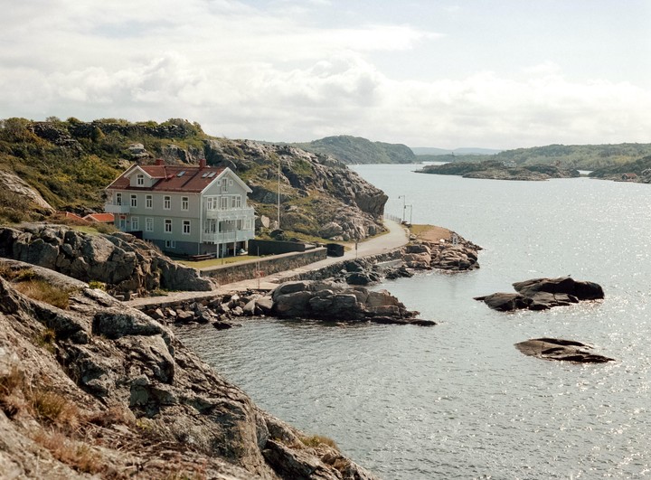 A large house with a red roof sits on a rocky coastline next to a body of water with several small islands.