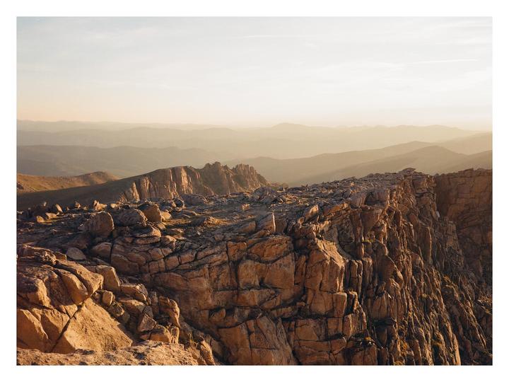 A rocky mountain ridge is illuminated by early morning light, with hazy mountains in the distance.
