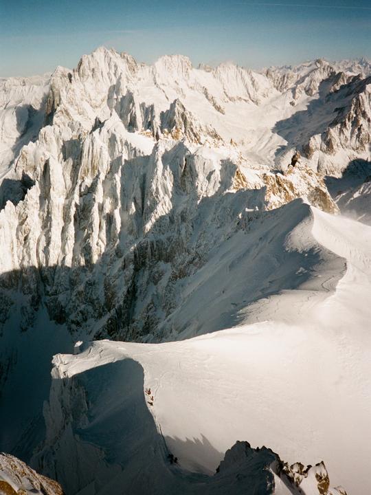 A group of people are climbing a snow-covered mountain ridge with a vast mountain range in the background.