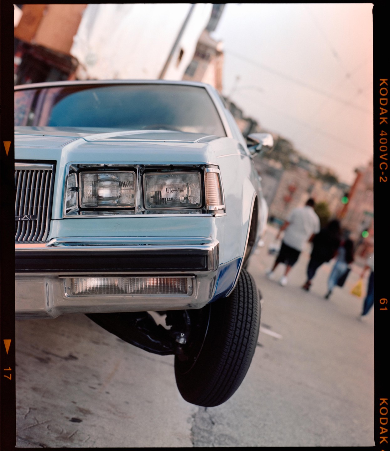 A light blue lowrider car is shown with its front wheel lifted off the ground on a street.