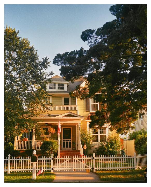 A yellow house with a white picket fence and an American flag hanging from the porch is surrounded by trees.