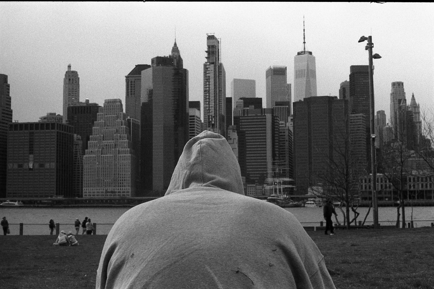 A person in a hooded sweatshirt looks out at the new york city skyline from across the water.
