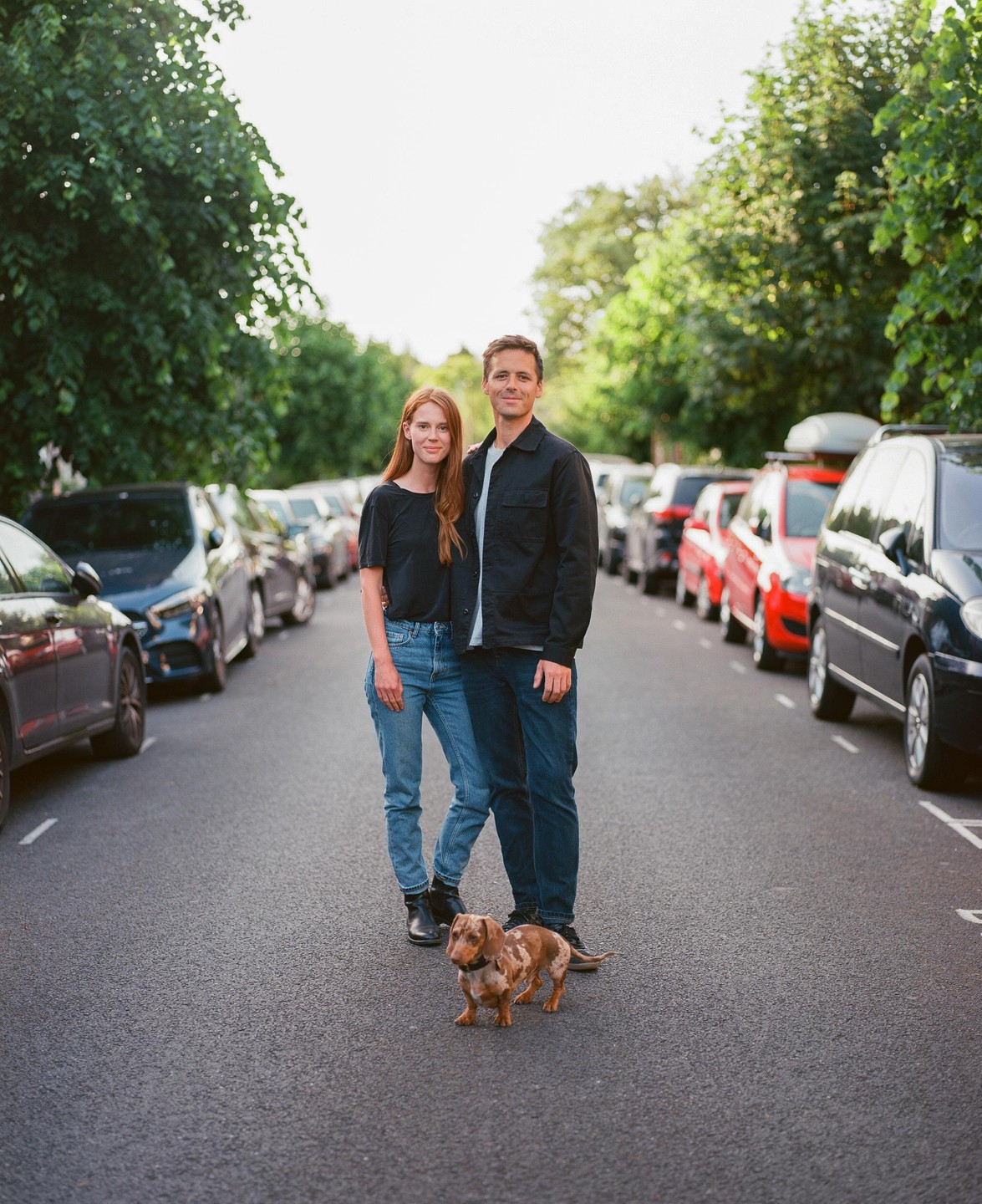 A couple stands with their dog in the middle of a street lined with parked cars.