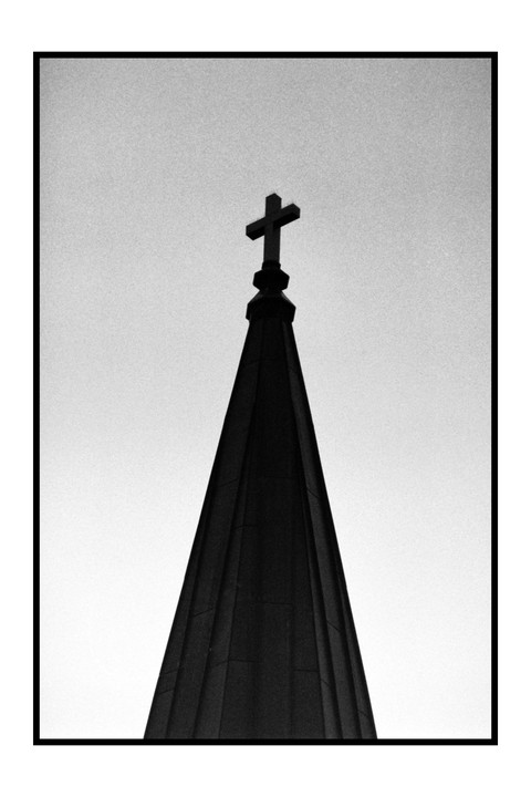 A cross sits atop a dark, pointed steeple against a bright sky.