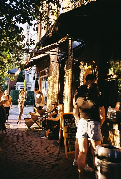 People are gathered on a brick sidewalk outside a building with an awning during golden hour.