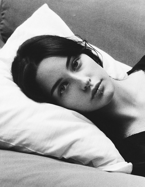 A woman with dark hair and a lip piercing lies on a pillow, looking directly at the viewer.