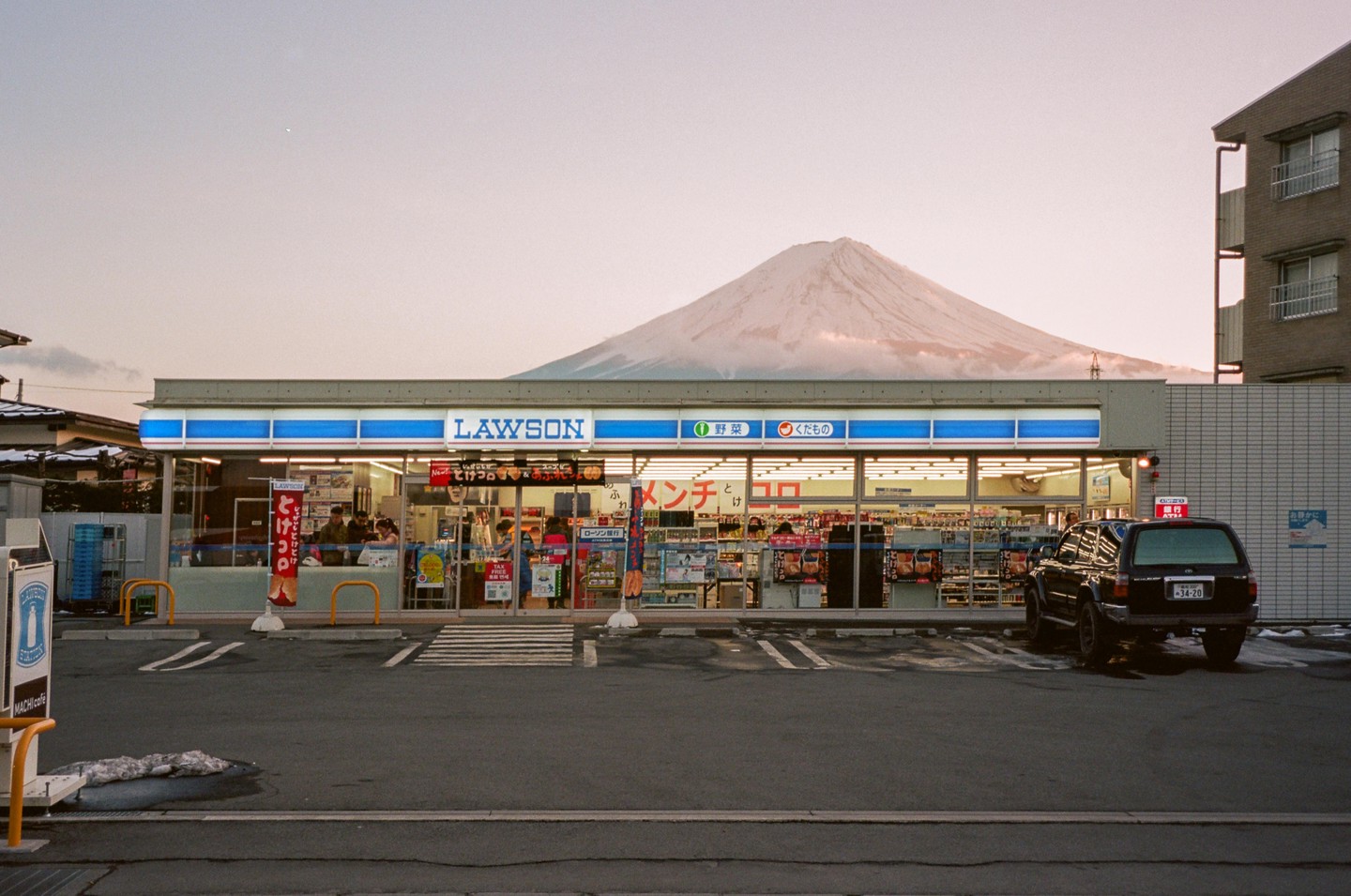 A Lawson convenience store with a snow-capped mountain in the background and a black vehicle parked in front.
