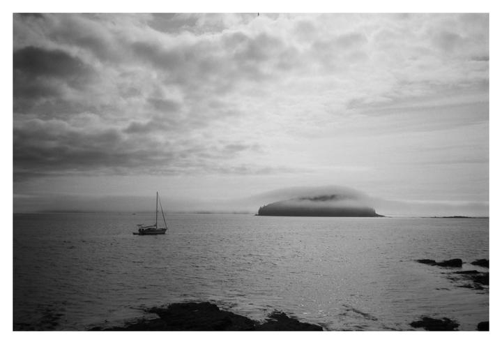 A sailboat floats on the water with a fog-covered island in the distance under a cloudy sky.
