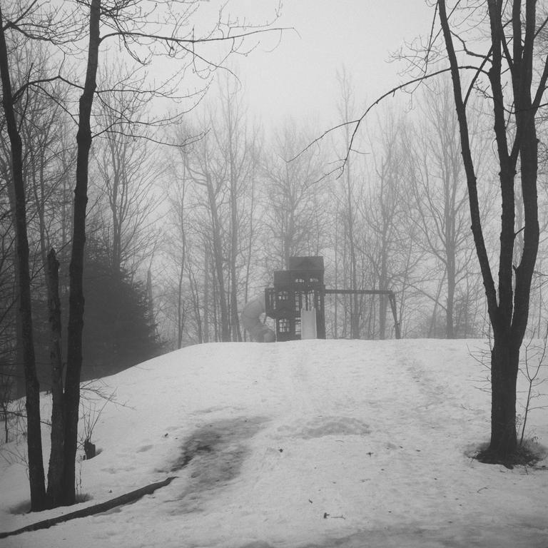 A playground structure with a slide and swings is visible on a snow-covered hill, surrounded by bare trees in a foggy landscape.