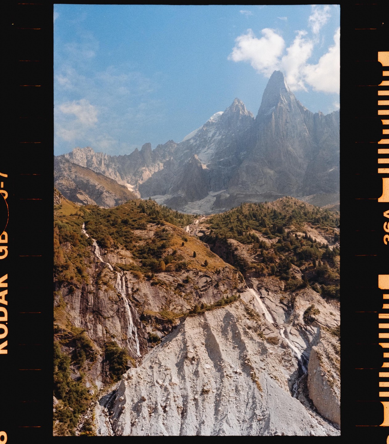 A waterfall cascades down a rocky mountain with a forest and jagged peaks under a blue sky.