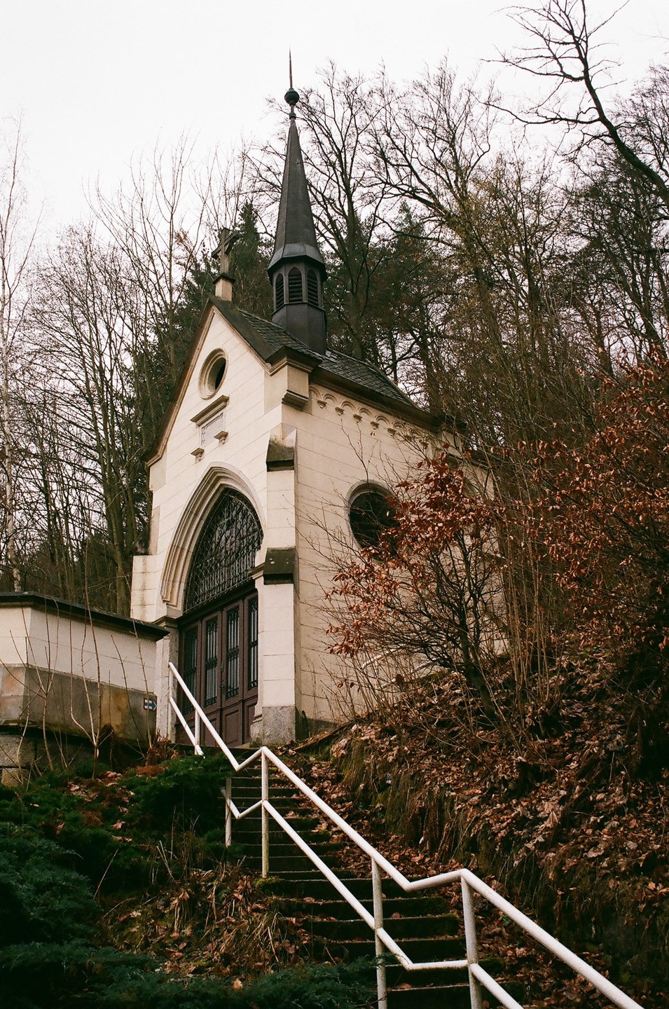 A small, light-colored church with a dark steeple stands on a hill, surrounded by bare trees and brown foliage, with a white railing leading up to its entrance.