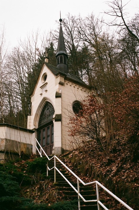 A small, light-colored church with a dark steeple stands on a hill, surrounded by bare trees and brown foliage, with a white railing leading up to its entrance.