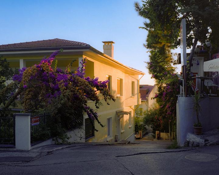 A street with a house covered in purple bougainvillea and a gelato shop sign is illuminated by golden hour light.