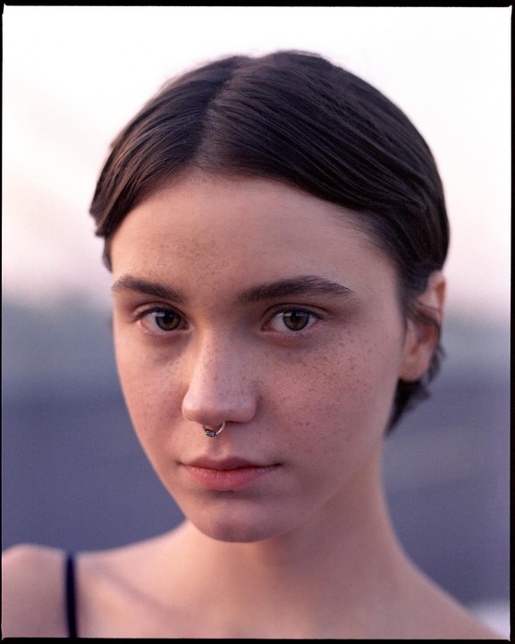 A young woman with short hair, freckles, and a septum piercing looks directly at the viewer.