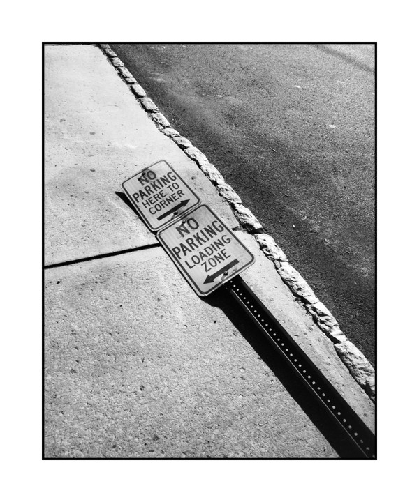 Two "no parking" signs are attached to a pole on a sidewalk next to a street.