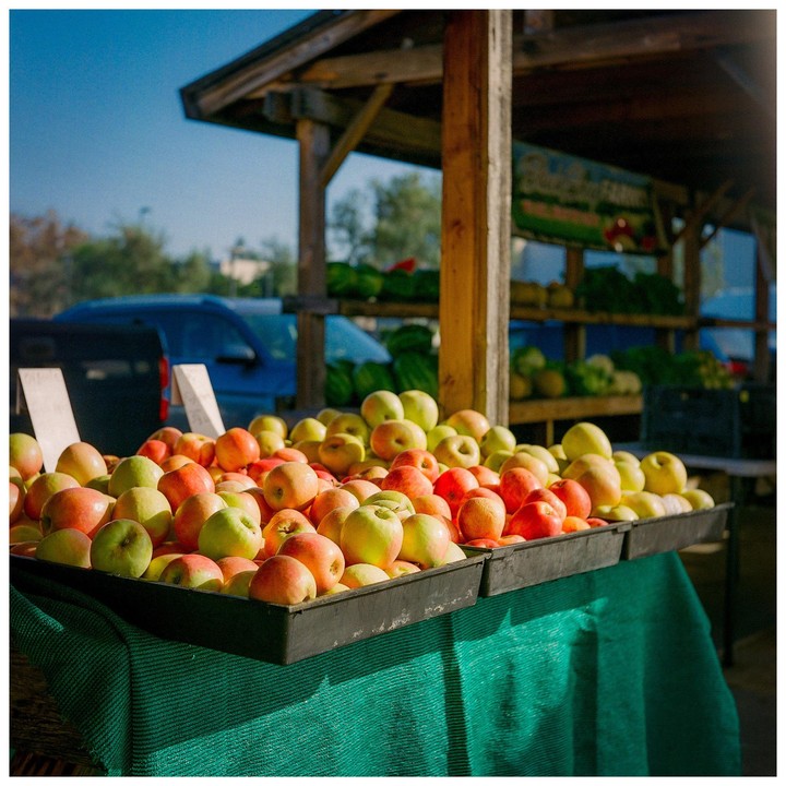 Trays of red and green apples sit on a table covered with a green cloth at an outdoor market stall.