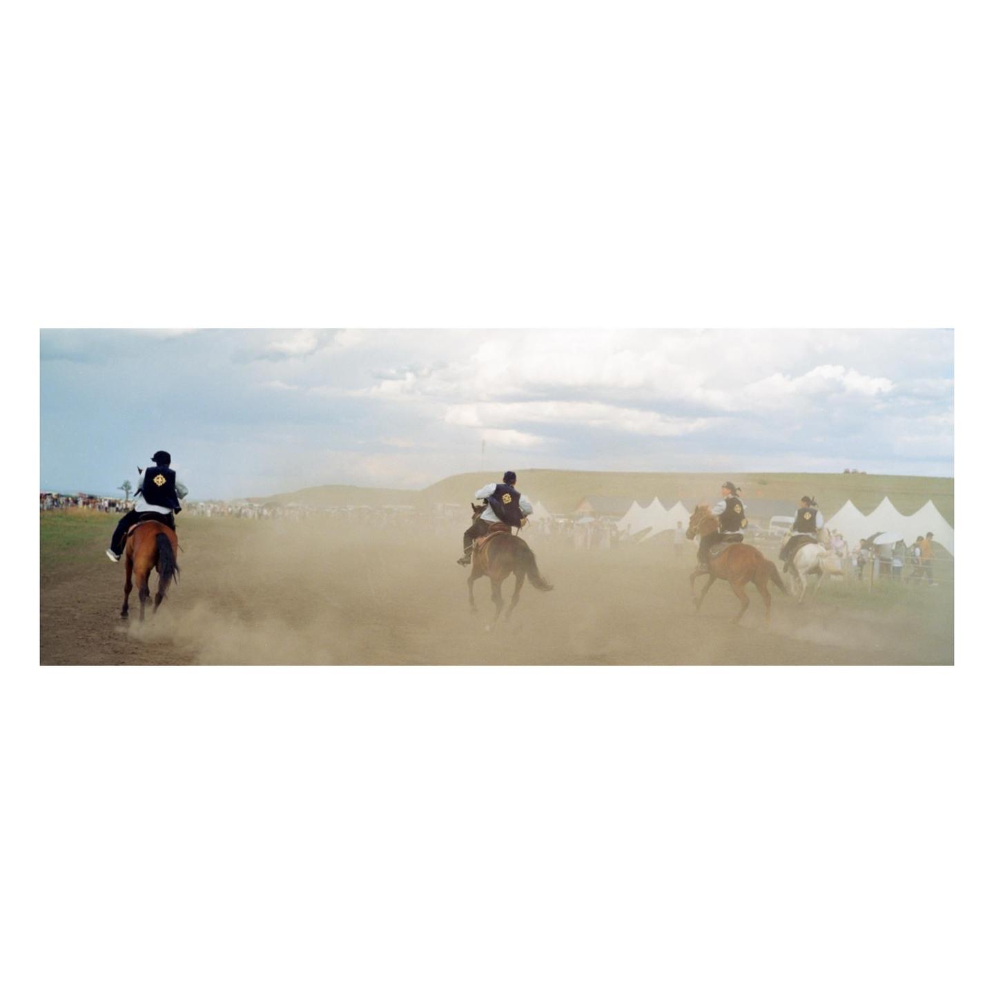 Several people on horseback race across a dusty field with white tents and hills in the background.