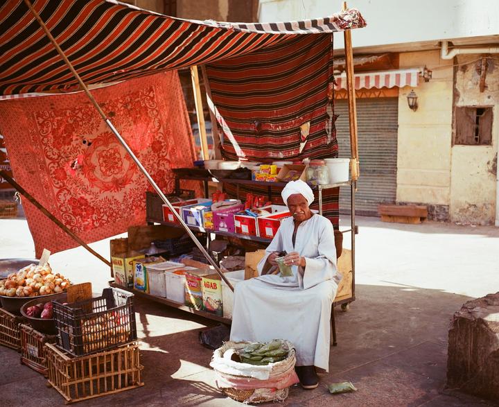 A man sits under a striped awning at a market stall with produce and packaged goods.