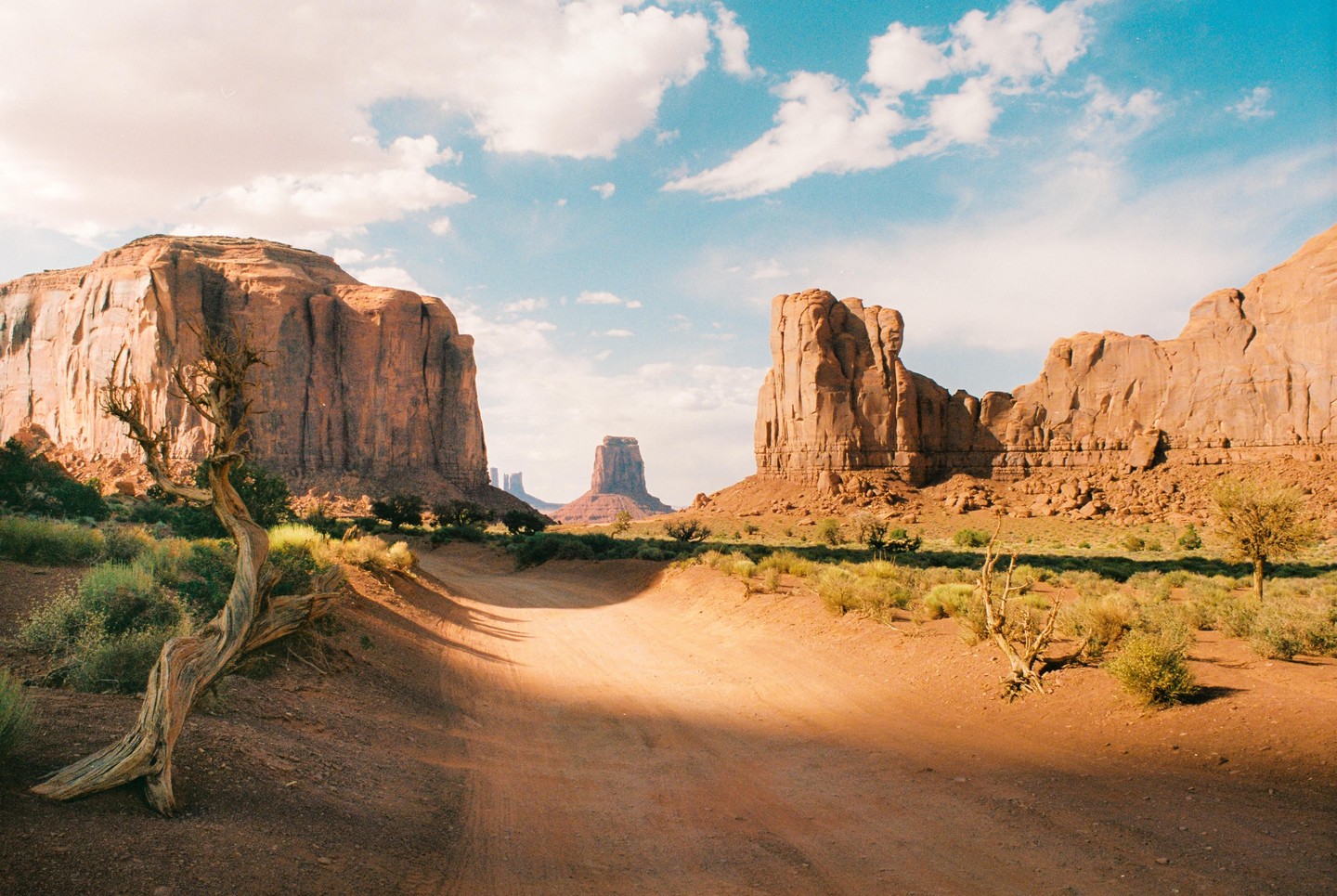 A dirt road leads through a desert landscape with large rock formations under a blue sky with clouds.