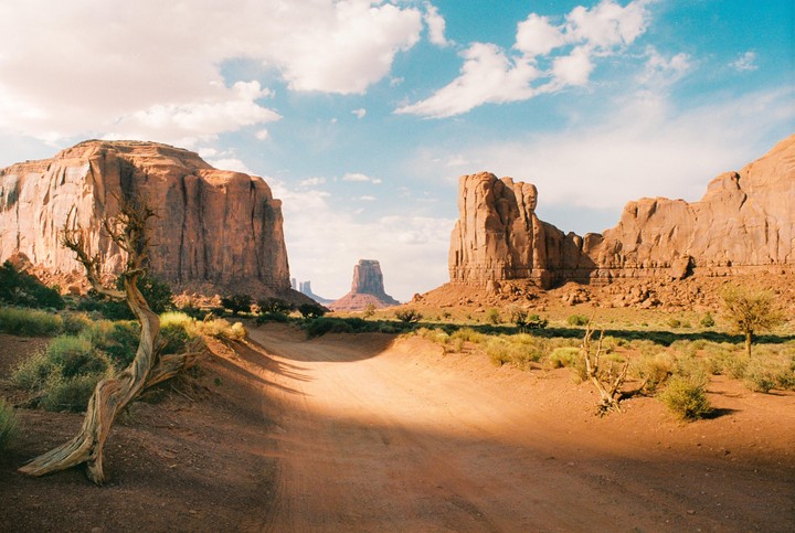 A dirt road leads through a desert landscape with large rock formations under a blue sky with clouds.
