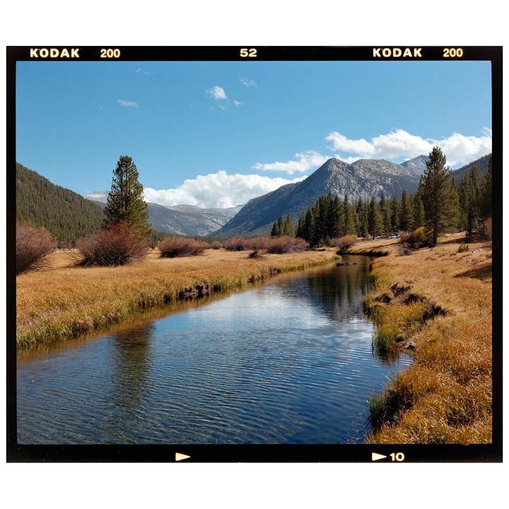 A river flows through a valley with mountains and trees under a blue sky with clouds.