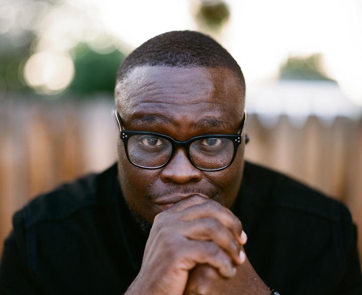 A man wearing glasses looks directly at the viewer with his hands clasped under his chin.