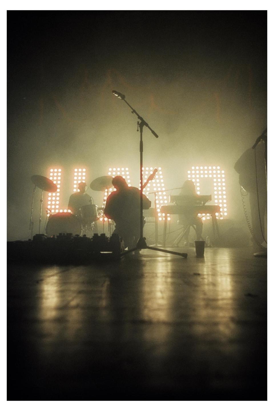 A band performs on a stage with bright lights spelling out a word in the background.