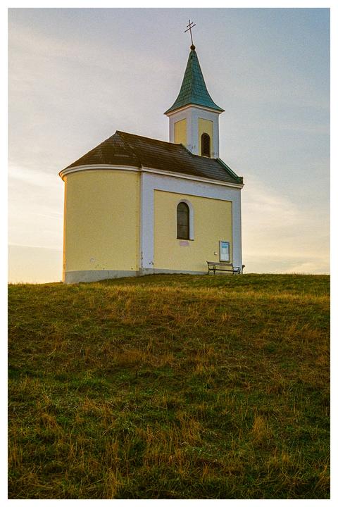 A small chapel with a green steeple stands on a grassy hill under a pale sky.