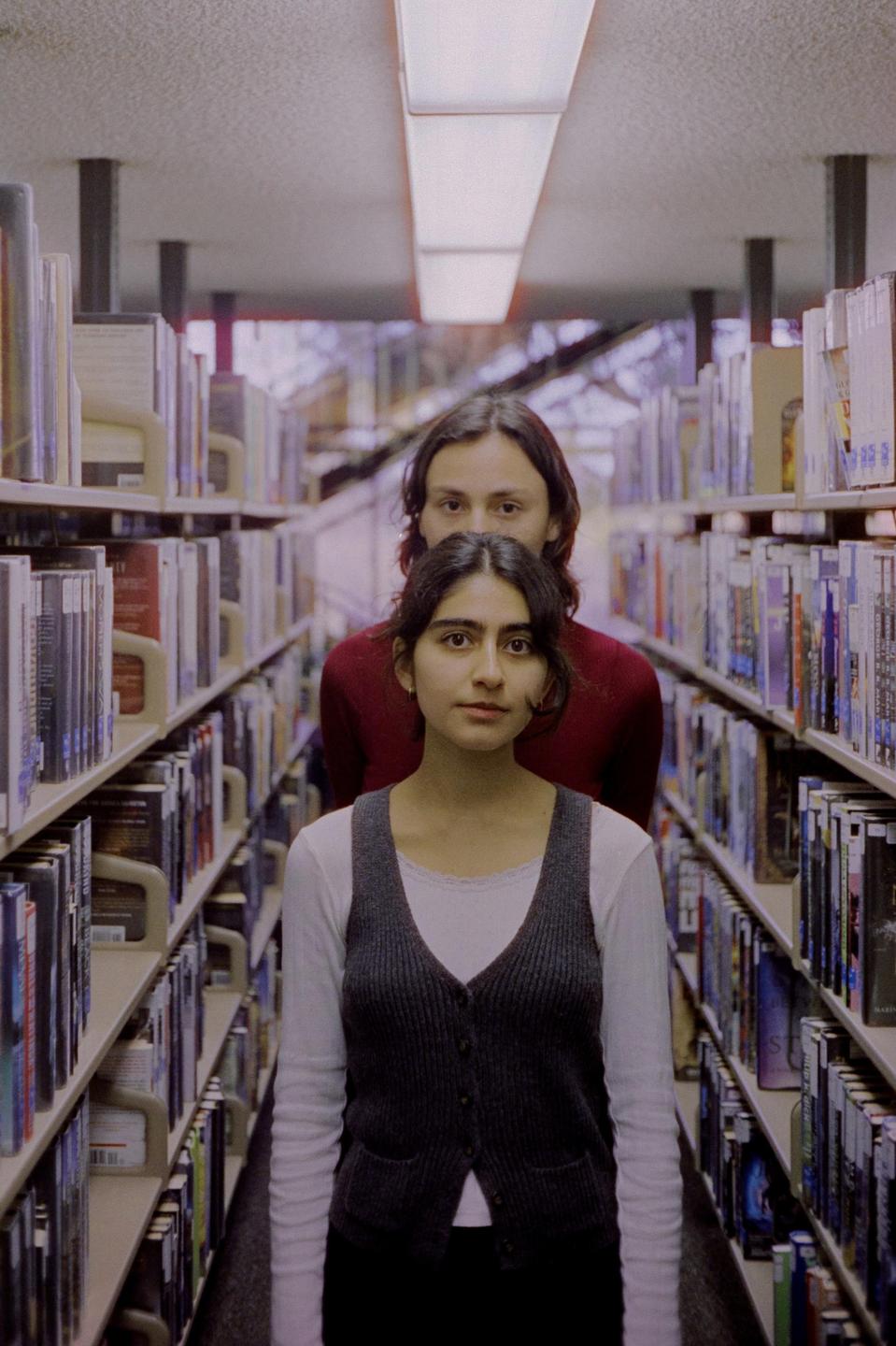 A woman stands in front of a man in a library aisle, both looking directly at the viewer.