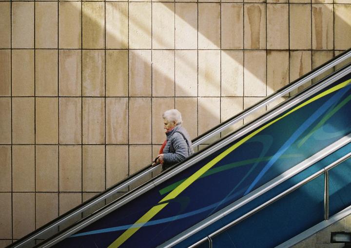 A woman rides an escalator next to a tiled wall with a diagonal beam of light.