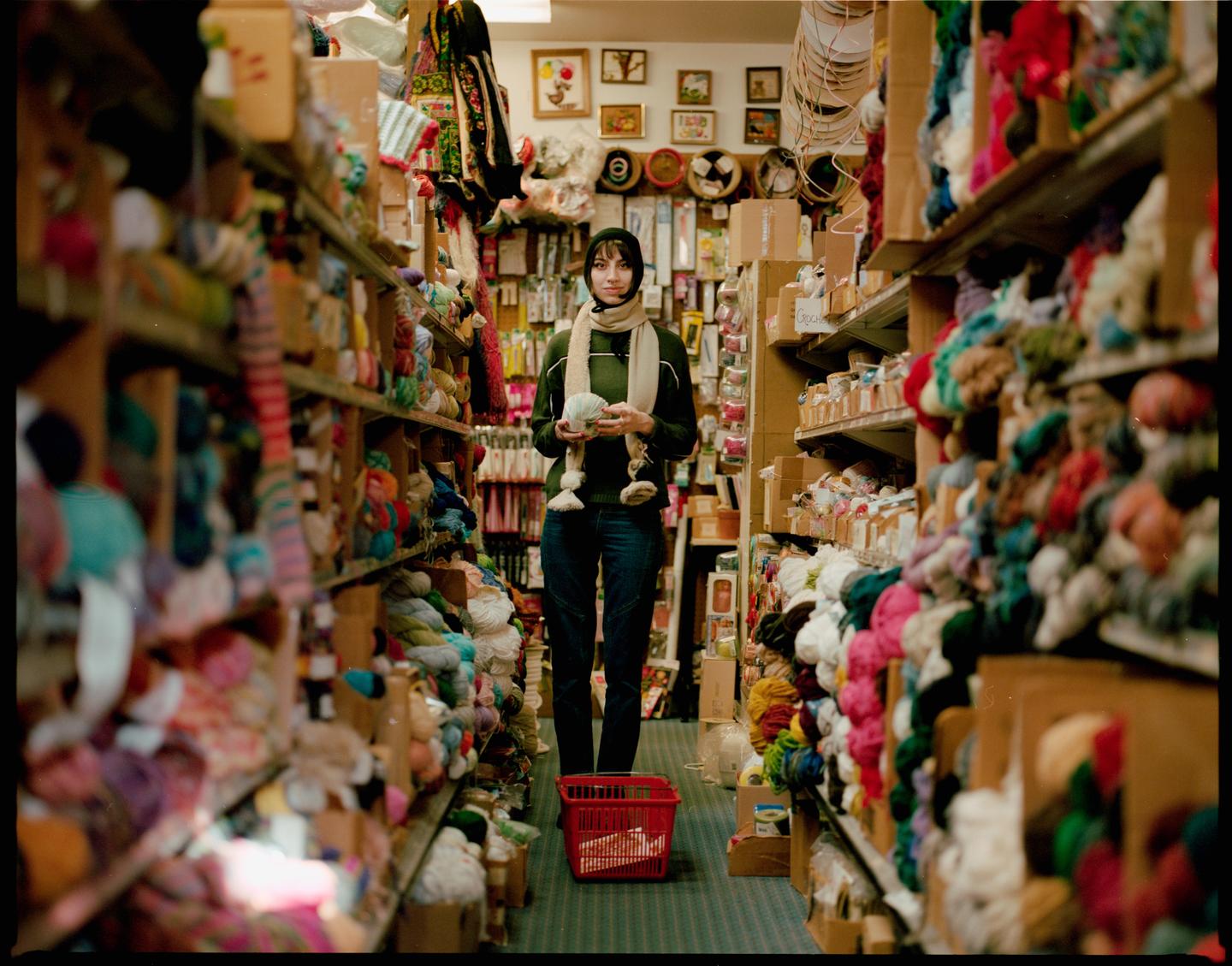 A woman stands in a narrow aisle of a yarn shop, holding a skein of yarn.