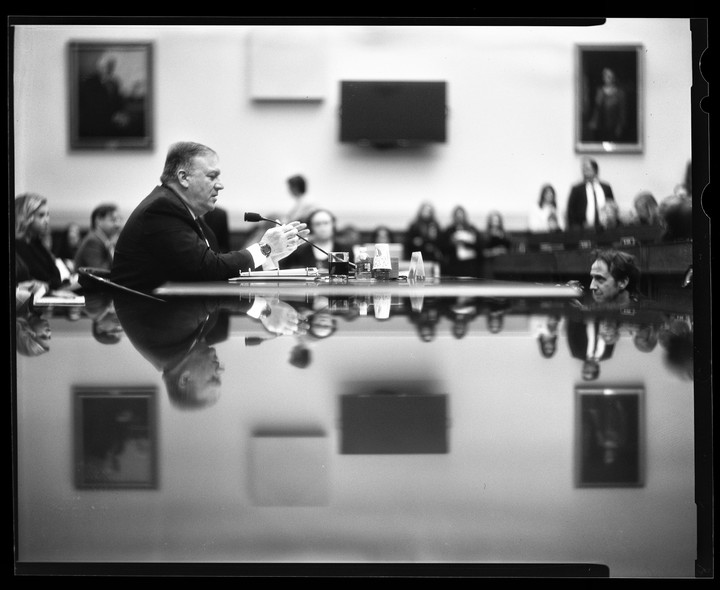 A man in a suit sits at a long table with a microphone, with his reflection visible on the table's surface.