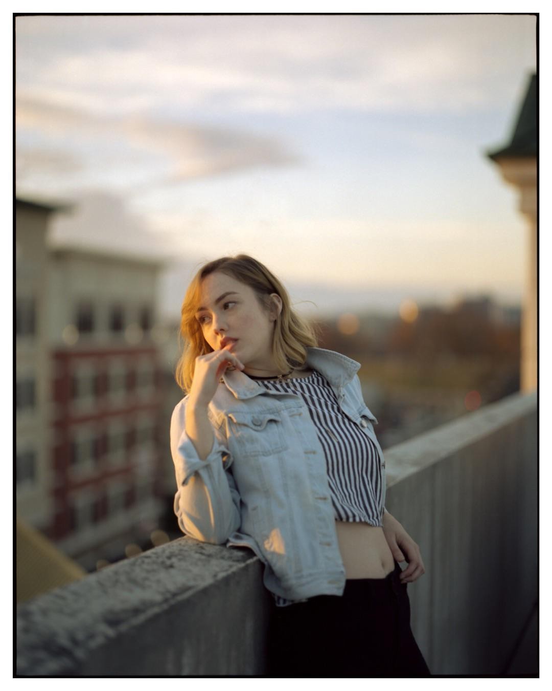 A woman in a denim jacket and striped top leans on a concrete ledge with a city in the background at sunset.
