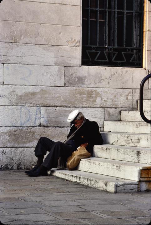 A man wearing a white cap and dark coat sits on stone steps next to a building with a barred window.