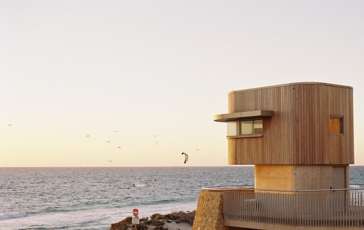 A wooden building with a balcony overlooks the ocean where a person is kitesurfing and birds are flying.