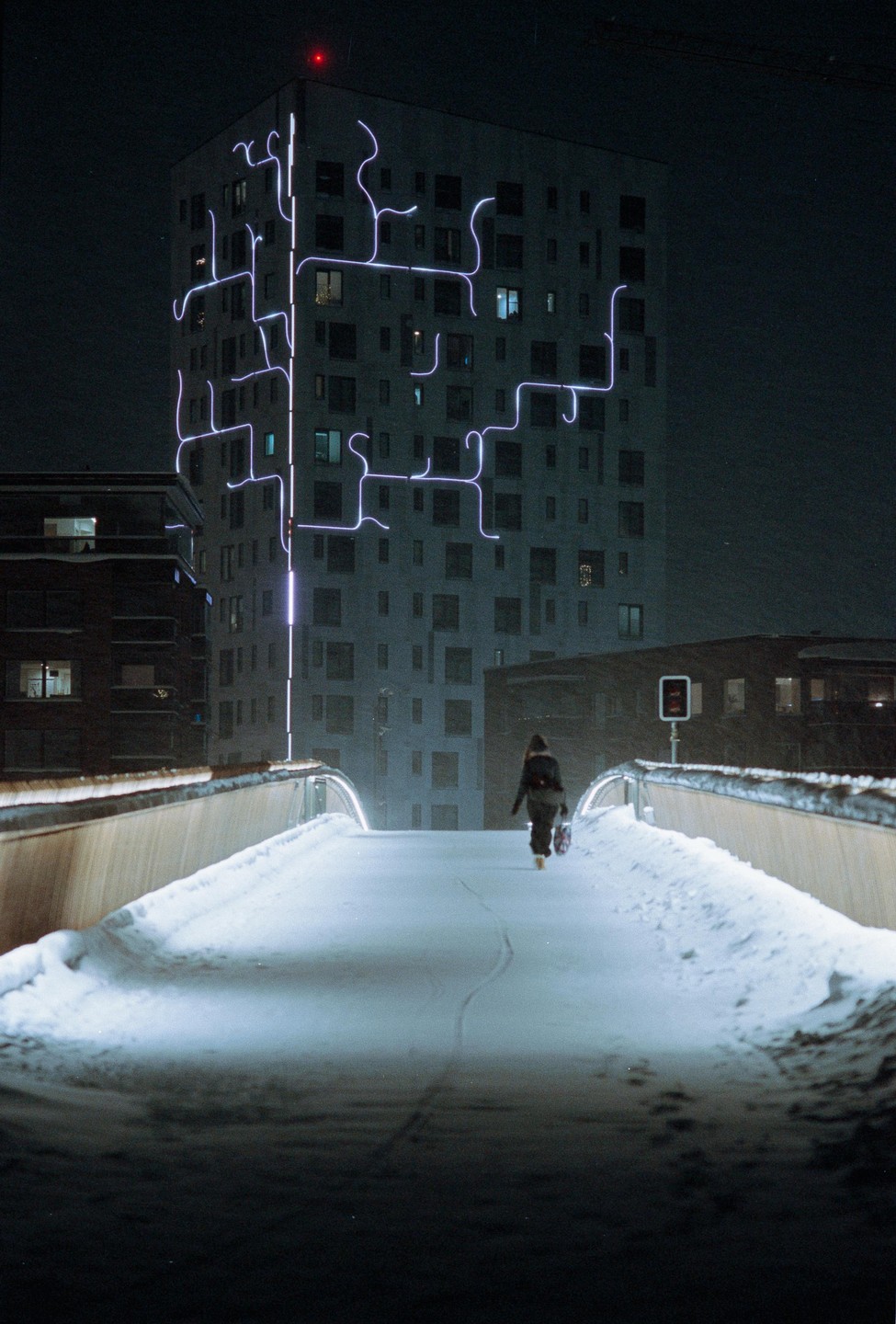 A person walks on a snow-covered bridge towards a building with glowing neon lights at night.