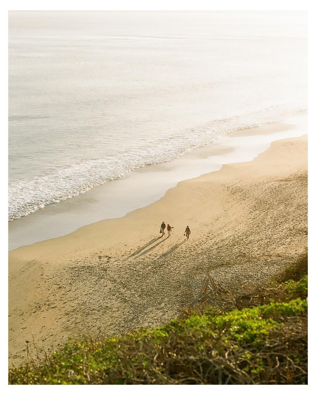 Three people walk along a sandy beach next to the ocean with long shadows stretching out before them.