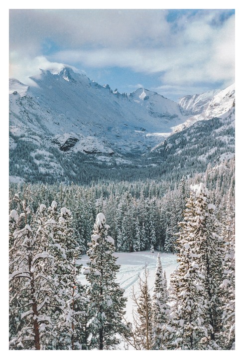 A snow-covered forest and valley are nestled between towering mountains under a cloudy sky.
