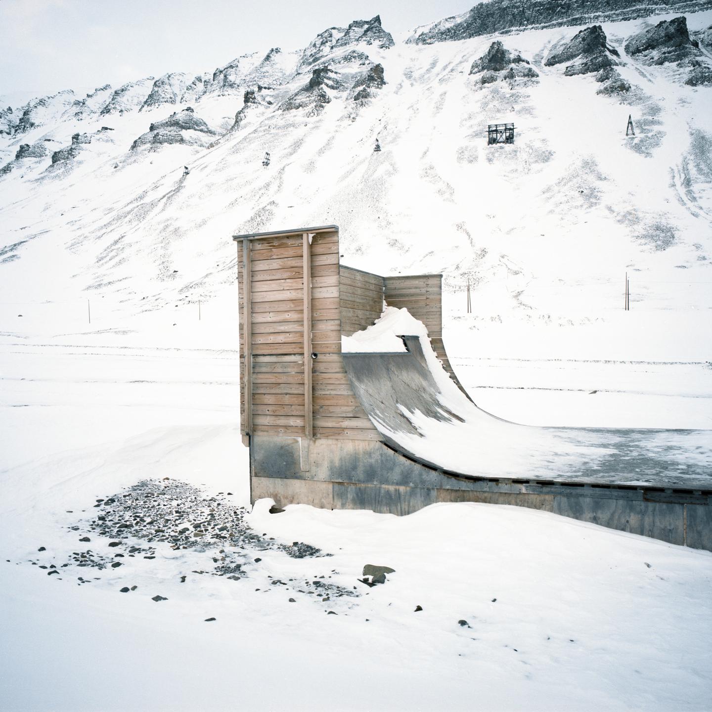 A wooden half-pipe covered in snow sits in a snowy landscape with mountains in the background.
