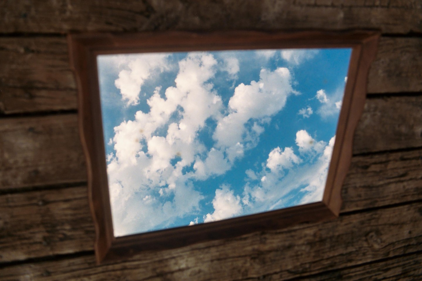 A wooden picture frame hangs on a wooden wall, reflecting a bright blue sky with white clouds.