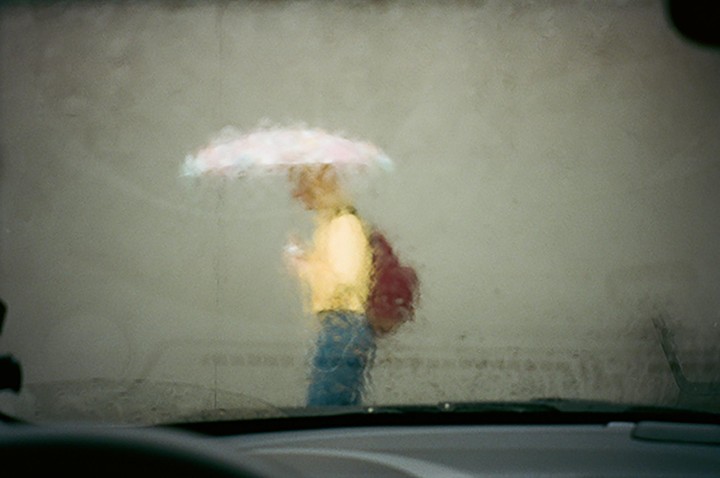 A person with an umbrella and backpack walks in the rain, viewed through a wet car windshield.