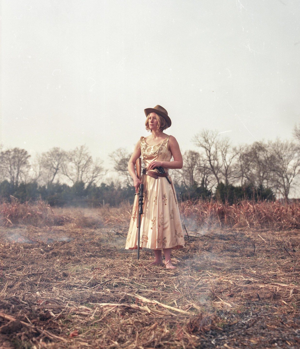 A woman in a dress and cowboy hat stands barefoot in a field holding a rifle and a pistol.